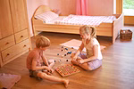 Zwei Kinder spielen mit dem Romanswerk Farbenspiel 10x10 und ordnen die 100 bunten Filzkugeln auf einem Holzboden an. Im Hintergrund sorgen ein Bett mit rosafarbener Bettwäsche und eine hölzerne Kommode für eine gemütliche Atmosphäre im Zimmer.