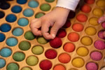Eine Kinderhand legt eine rote Filzkugel in das "Farbenspiel 10x10 inkl. 100 farbige Kugeln aus Filz" von Romanswerk das ein Holzbrett mit bunten Filzkugeln füllt die in kreisförmigen Schlitzen angeordnet sind.