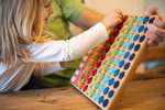 Ein Kleinkind steckt eine bunte Filzkugel in das mit bunten Löchern gefüllte Romanswerk Farbenspiel 10x10 während ein Erwachsener das Holzspielzeug auf einem Tisch festhält.