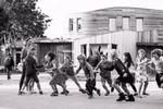Kinder spielen draußen auf dem Bürgersteig während ein Erwachsener zuschaut mit modernen Holzgebäuden im Hintergrund. Das Schwarz-Weiß-Bild erscheint in "Einblicke weltweit - Einblicke weltweit 100 Jahre Rudolf Steiner Pädagogik" vom Kehrer Verlag.