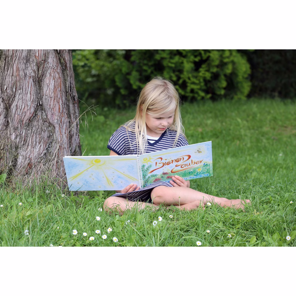 DUO Verlag Bienenzauber Mitmachbilderbuch_582 Ein junges Mädchen sitzt an einem sonnigen Tag im Gras unter einem Baum und liest das "Bienenzauber Mitmachbilderbuch" des DUO Verlags umgeben von Grün und kleinen weißen Blumen tief konzentriert auf das Buch.