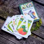 Ein Stapel illustrierter ABC-Spielkarten mit deutschen Wörtern liegt auf einer hölzernen Unterlage neben der ABC-Kartenspiel Lernspiel-Box des Waldow Verlags die mit farbenfrohen Motiven und dem Produkttitel versehen ist.