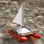 Der SUNNYSUE Bausatz-Katamaran (für 5-8 Jahre) schwimmt mit seinem weißen Segel und den roten Pontons auf klarem flachem Wasser unter dem man Kieselsteine sehen kann.