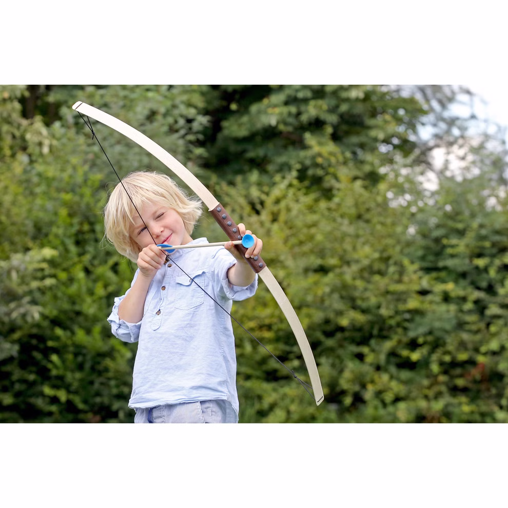 Spielzeugmanufaktur VAH Pfeil und Bogen L 100 cm_1335 Ein zielstrebiges Kind im hellblauen Hemd zielt mit der Spielzeugmanufaktur VAH "Pfeil und Bogen L 100 cm" im Freien vor grünen Büschen und Bäumen stehend die blonden Haare nach hinten gezogen bereit den Pfeil abzuschießen.