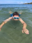Ein Junge trägt die Schildkröt Junior Schwimmbrille Bali 4+ blau mit Aufbewahrungstasche schwimmt im flachen grünlichen Wasser in der Nähe eines Strandes das Gesicht teilweise über der Wasseroberfläche und die Arme ausgebreitet unter einem blauen Himmel mi