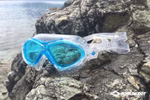 Blaue Junior-Schwimmbrille "Bali 4+" von Schildkröt mit Aufbewahrungstasche ruht auf Felsen in der Nähe von ruhigem Wasser. Die Marke Schildkröt ist in der unteren rechten Ecke sichtbar.