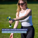 Eine lächelnde Frau mit Sonnenbrille weißem Tanktop und schwarzer Hose spielt mit dem Schildkröt Neopren Handfederball Peteca auf dem Rasen. Text in Deutsch und Englisch hebt die weiche griffige Neoprenhülle hervor.