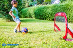 Schildkröt Fußballtore Pop-Up mit Aufbewahrungstasche _26497 Ein kleiner Junge in Grün kickt einen bunten Ball in Richtung des Schildkröt Fußballtore Pop-Up mit Aufbewahrungstasche auf einer Wiese mit Bäumen und Büschen dahinter. In der Ecke ist das Schildkröt-Logo zu sehen.
