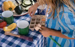 Ein Kind in einem blau gestreiften Hemd füllt ein kleines Setzlingstablett mit Erde an einem mit einer blau karierten Tischdecke bedeckten Tisch umgeben von Gemüse Bechern und dem 6-teiligen Set Grapat Tools von Grapat.