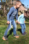 Jamso Design Shaddy Hufe _26707 Zwei Kinder spielen im Gras in der Nähe eines Hauses und von Bäumen. Das ältere Kind läuft auf Shaddy-Hufe-Holzstelzen mit Seilen von Jamso Design während das jüngere Kind zusieht und in der Nähe läuft. Beide tragen Jeans und langärmelige Hemden.