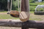 Jamso Design Shaddy Hufe _26709 Zwei hölzerne Hufschuhe von Jamso Design Shaddy Hufe hängen an Seilen an einem Holzzaun. Ein weißes Pferd grast im unscharfen Hintergrund in dem grünes Gras und Bäume zu sehen sind.