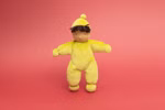 Die Käthe Kruse Waldorfpuppe Lolo moos ist eine weiche Plüschpuppe mit hellbrauner Haut schwarzem Garnhaar und einem gelben Outfit mit passender Mütze posiert vor einem einfarbigen korallenrosa Hintergrund.