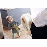 Ein lächelndes Kleinkind hält die Käthe Kruse Waldorfpuppe Lolo moos während es barfuß auf einem Holzboden in einer modernen Küche steht. In der Nähe sitzt ein Erwachsener auf einem Stuhl mit einem flauschigen weißen Bezug.