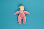 Die Käthe Kruse Waldorfpuppe Lolo Rose 21 cm ist eine weiche Plüschpuppe mit heller Haut blondem Garnhaar und einem rosa Outfit mit Hut die auf einem einfarbig blauen Hintergrund liegt.