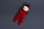 Die Käthe Kruse Waldorfpuppe Lolo Rubin 21 cm ist eine weiche Puppe mit einem beigen Gesicht weißen Haaren einfachen Augen und einem roten Plüsch-Outfit mit Hut die auf einem dunkelgrauen Hintergrund liegt.