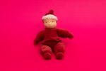 Die Käthe Kruse Waldorfpuppe Lolo rubin (21 cm) eine Plüschpuppe mit hellbeigem Gesicht weißem Haar rotem Outfit und Hut ist vor einem hellrosa Hintergrund sitzend abgebildet.