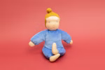 Die Käthe Kruse Waldorfpuppe Nana wolke 35 cm ist eine weiche Stoffpuppe mit gelber Strickmütze blauem Outfit und hellem Haar die im Schneidersitz auf einem einfarbig roten Hintergrund sitzt.
