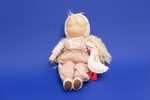 Die Käthe Kruse Waldorfpuppe Hannah 38 cm eine Stoffpuppe mit blondem Garnhaar Kopftuch rosa Latzhose und gepunktetem Hemd sitzt vor einem blauen Hintergrund und hält eine kleine weiße Stoffgans.