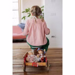 Ein kleines Kind mit geflochtenem Haar sitzt auf einem kleinen Holzwagen der mit der Käthe Kruse Waldorfpuppe Hannah (38 cm) gefüllt ist und blickt in ein gemütliches sonnendurchflutetes Zimmer mit Holzboden und einem rosa Sofa im Hintergrund.