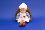 Die Käthe Kruse Waldorfpuppe Tilda 38 cm eine handgefertigte Stoffpuppe mit braunem gelocktem Haar blauer Haube blauem Kleid mit weißer Schürze und rotem Hühnermotiv und roten Schuhen wird vor einem einfarbig blauen Hintergrund gezeigt.