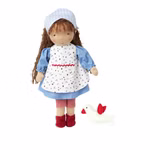Die Käthe Kruse Waldorfpuppe Tilda 38 cm mit braunen Locken blauem Kleid weißer Schürze mit roter Borte roten Stiefeln und blaukariertem Kopftuch steht neben einem kleinen weißen Plüschvogel mit roten Flügeln und Schnabel.