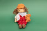 Die Käthe Kruse Waldorfpuppe Toni 38 cm sitzt auf grünem Hintergrund hat braunes Garnhaar eine gelbe Mütze eine rote Latzhose und einen weiß gemusterten Schal und hält ein kleines braunes Eichhörnchen-Plüschtier.