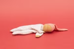 Die Käthe Kruse Waldorfpuppe Nini Liebchen 21 cm ist eine weiche Stoffpuppe mit gesticktem Gesicht bekleidet mit einem weißen Outfit mit roten Punkten und einem langen rosa Hut die auf der Seite liegend vor einem einfarbig roten Hintergrund abgebildet ist.