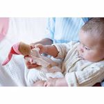 Ein leicht bekleidetes Baby liegt auf dem Schoß und betrachtet die Käthe Kruse Waldorfpuppe Nini Liebchen 21 cm - eine kleine Puppe in Weiß mit roten Punkten und einem rosa Hut.