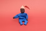 Die Waldorfpuppe Nini von Käthe Kruse (Sommerwolke 24 cm) ist eine weiche Stoffpuppe mit dunkelbrauner Haut gekleidet in Blau mit rosa Zipfelmütze die auf einem einfarbig roten Hintergrund sitzt.