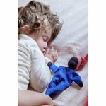 Ein kleines Kind schläft auf der Seite die Augen geschlossen und hält die Käthe Kruse Waldorfpuppe Nini Sommerwolke 24 cm mit blauem Kleid und rotem Hut dicht auf einer hellen Decke.