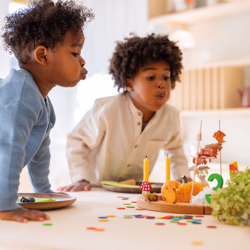 Grimm's Konfettitaler Regenbogen aus Holz _28819 Zwei kleine Kinder blasen Kerzen auf einem Geburtstagskuchen aus der an einem mit Grimms Konfettitaler Regenbogen aus Holz geschmückten Tisch steht und schaffen so eine farbenfrohe festliche Atmosphäre in einem hellen fröhlichen Raum.