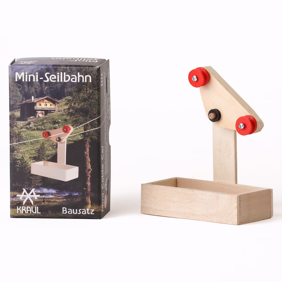Kraul Mini-Seilbahn Wagen mit 5 m Seil Bausatz _29537 Ein Kraul Mini-Seilbahn Wagen mit 5 m Seil Bausatz steht neben einem kleinen hölzernen Seilbahnmodell mit roten Rädern; auf der Schachtel sind eine Berghütte und eine Seilbahn abgebildet beide vor einem weißen Hintergrund.