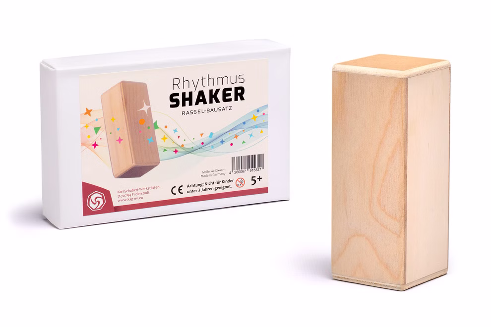 Karl Schubert Gemeinschaft Bausatz Rhythmus Shaker Klangspielzeug_29970 Ein rechteckiger Bausatz Rhythmus Shaker Klangspielzeug aus Holz von Karl Schubert Gemeinschaft steht neben seiner weißen Schachtel mit bunten Noten und Sternen beschriftet für Kinder ab 5 Jahren.