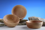 Karl Schubert Gemeinschaft Holzschale aus Esche _32960 Drei leichte Holzschalen aus Esche von der Karl Schubert Gemeinschaft werden auf einer neutralen Fläche mit zwei flachen Steinen vor einem blauen Hintergrund mit Farbverlauf präsentiert.