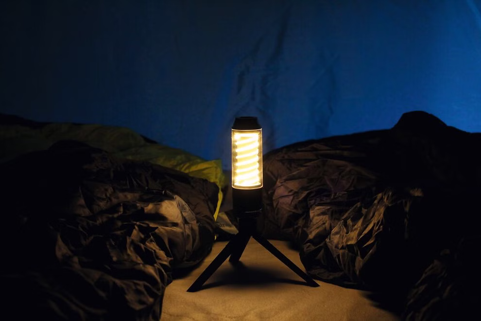 moses. Verlag Expedition Natur Outdoor Allroundlicht _29957 Die Expedition Natur Outdoor Allroundlicht vom moses. Verlag steht auf sandigem Boden inmitten zerknüllter dunkler Schlafsäcke im Hintergrund eine blaue Zeltwand.