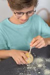 Ein Kind mit Brille und hellblauem Hemd gräbt mit einem Holzstab ein Set Mondsteine zum Ausgraben aus dem moses. Verlag mit Pulver auf dem Tisch verstreut.