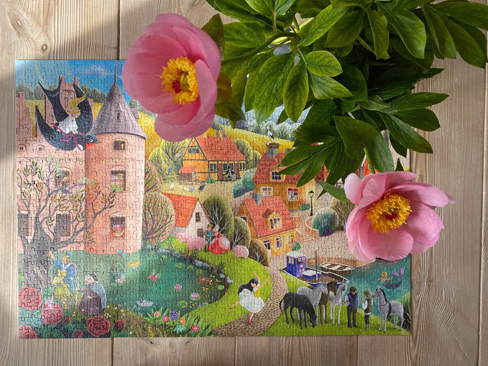 Penny Puzzle Puzzle Home of Fairytales 1000 Teile _30091 Ein fertiges Penny Puzzle "Home of Fairytales" 1000 Teile zeigt ein lebendiges Dorf mit Häusern Menschen und Tieren auf einer Holzoberfläche. Zwei rosa Pfingstrosen und grüne Blätter bedecken teilweise die obere rechte Ecke.
