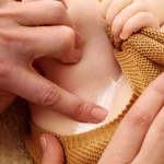 Weleda Calendula Wundschutzcreme parfümfrei 75 ml _31068 Ein Erwachsener trägt die Weleda Calendula Wundschutzcreme parfümfrei 75 ml eine weiße Creme auf die Haut des Babys im Windelbereich auf während das Baby ein braunes Strickkleidungsstück trägt.