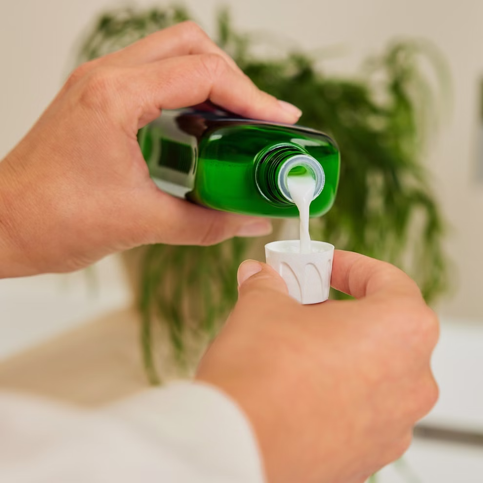 Weleda Weleda Edeltannen-Erholungsbad 200 ml _30866 Jemand gießt eine weiße Flüssigkeit aus einer grünen Weleda Edeltannen-Erholungsbad 200 ml Flasche in einen kleinen Plastikbecher wobei eine Blattpflanze im Hintergrund verschwimmt.