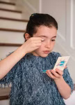 Ein junges Mädchen in einem blauen Oberteil benutzt die Namaki Cosmetics Glitzer Kosmetiktasche Blauer Fuchs Lidschattenpalette und Glitzer Lipgloss und trägt Make-up auf während sie in einen kleinen Handspiegel in der Nähe einer Treppe im Haus schaut.