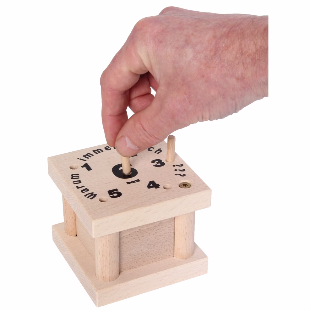 Bartl Warum immer ich? Würfelspiel_31722 Eine Hand hebt einen Holzpflock aus dem Bartl "Warum immer ich?" Würfelspiel - eine quadratische Schachtel mit nummerierten Löchern und Text auf der Oberseite die an ein Puzzle oder Lernspiel erinnert.