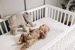 Senger Kuscheltier Elefant groß Wärmekissen_32013 Ein Baby in einem gemusterten Schlafanzug liegt auf einer weißen Kinderbettmatratze und blickt auf das Senger Kuscheltier Elefant groß Wärmekissen das neben ihm im Kinderbett liegt.
