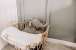 Senger Kuscheltier Elefant groß Wärmekissen_32014 In einer Ecke steht ein Weidenkorb auf einem Teppich mit einem Senger Kuscheltier Elefant groß Wärmekissen und einer gefalteten weißen Decke vor hellgrünen und weißen Wänden.