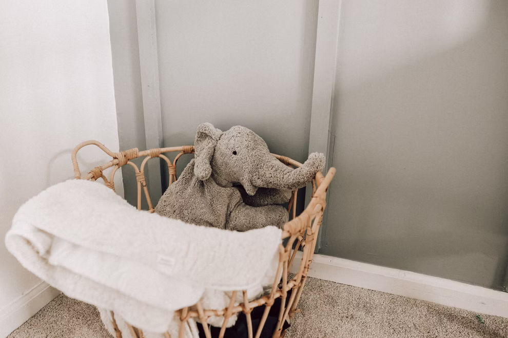 Senger Kuscheltier Elefant groß Wärmekissen_32014 In einer Ecke steht ein Weidenkorb auf einem Teppich mit einem Senger Kuscheltier Elefant groß Wärmekissen und einer gefalteten weißen Decke vor hellgrünen und weißen Wänden.