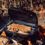 Der Stabilotherm Lagerfeuer Grilleisen grillt ein Gemüse- Grünzeug- und Käsesandwich über einem offenen Lagerfeuer wobei die Holzscheite und Flammen im Freien sichtbar sind.
