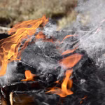 Das Stabilotherm Lagerfeuer Grilleisen eine schwarze gusseiserne Pfanne wird über einem Lagerfeuer im Freien erhitzt. Leuchtend orangefarbene Flammen und Rauch steigen um sie herum auf während sie auf einem Metallrost ruht.
