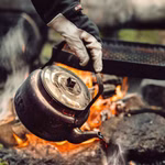 Eine behandschuhte Hand gießt Flüssigkeit aus dem Stabilotherm Kaffeekessel für das Lagerfeuer 2 l neben einem Lagerfeuer mit brennendem Holz und Steinen ein und fängt so die Essenz des Kaffee- oder Teebrühens im Freien ein.