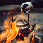 Eine behandschuhte Hand hält den Griff eines Stabilotherm Kaffeekessels für das Lagerfeuer 2 l der über einem offenen Lagerfeuer mit leuchtend orangefarbenen Flammen um ihn herum auf brennenden Holzscheiten erhitzt wird.