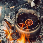 Der Stabilotherm Kaffeekessel für das Lagerfeuer 2 l sitzt über offener Flamme gefüllt mit dunkler Flüssigkeit und Orangenscheiben während eine behandschuhte Hand den Griff inmitten der Glut festhält.