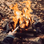 Eine Person benutzt den Stabilotherm Teleskop Grillstab um Essen über einem Lagerfeuer zu rösten mit Steinen und brennenden Holzscheiten umgeben von verstreuten Blättern im Hintergrund.